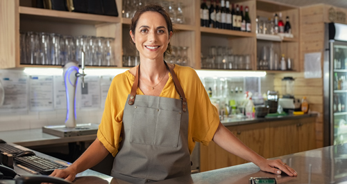 happy smiling woman restaurant owner stood at counter bar in yellow shirt and grey apron