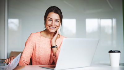 smiling woman with dark brown hair in peach top sat at white laptop
