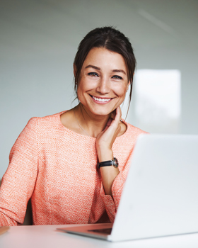 smiling woman in peach coloured top and black watch sat at desk on laptop