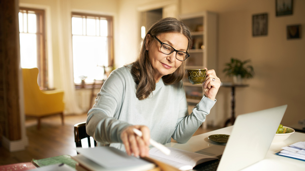 elder woman in grey long sleeve top black glasses drinking tea working at table in home on silver laptop